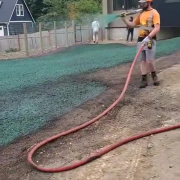 Worker spraying hydroseed on residential lawn area near wooden fence and custom home.