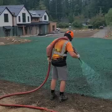 Worker applying blue-green hydroseed spray on bare soil near modern custom home in Snoqualmie.
