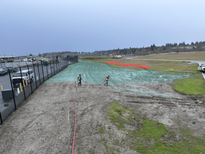 Workers spraying liquid on grass near parking lot and racetrack.