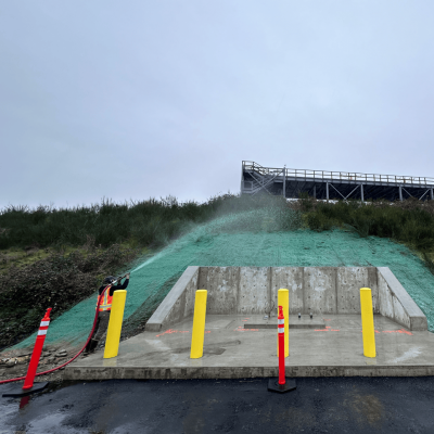 Worker applying green hydroseed to slope near concrete structure and yellow bollards.
