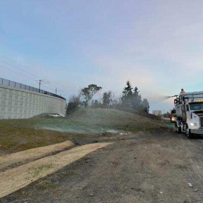 Truck spraying erosion control product on roadside slope near highway retaining wall.