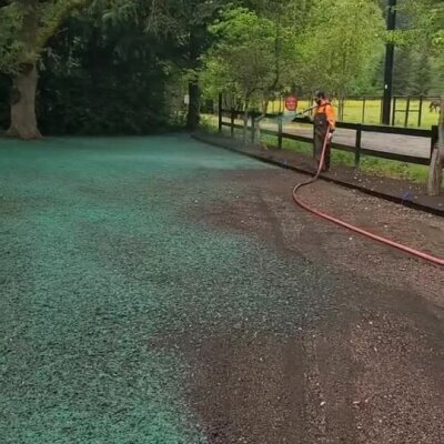 Hydroseeding worker spraying green grass seed slurry on large outdoor ground area.