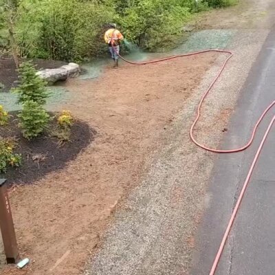 Worker spraying hydroseed on prepared soil along roadside near landscaped area.