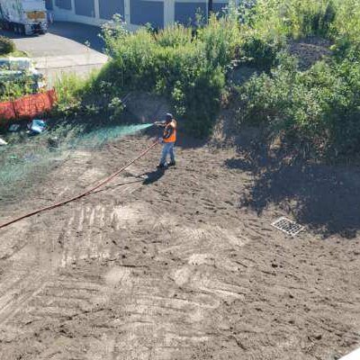 Worker applying hydroseed on sloped soil area near industrial facility in Seattle.