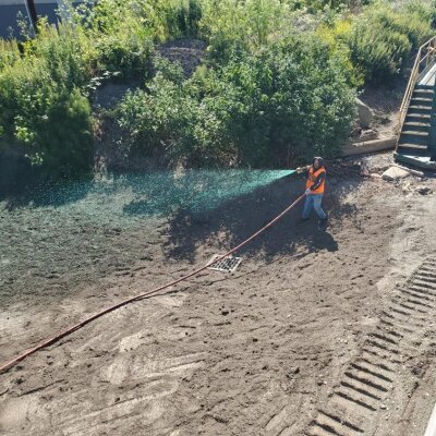 Worker applying green hydroseeding mixture on soil near vegetation and equipment.