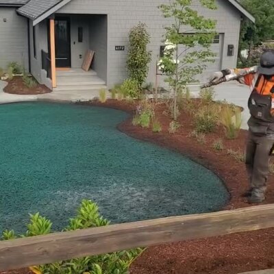 Worker spraying hydroseed on residential lawn with mulch and newly planted shrubs.