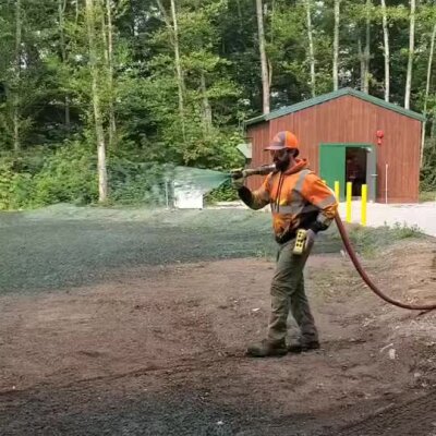 Worker spraying erosion control mulch near a building in wooded area.