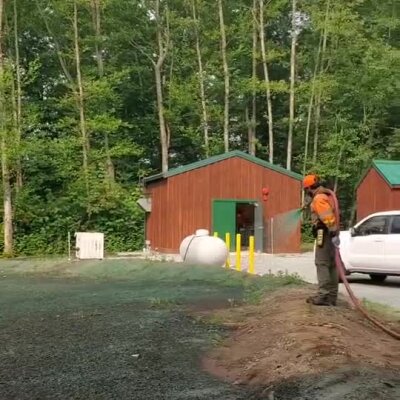 Worker spraying erosion control mulch near animal shelter buildings in wooded area.