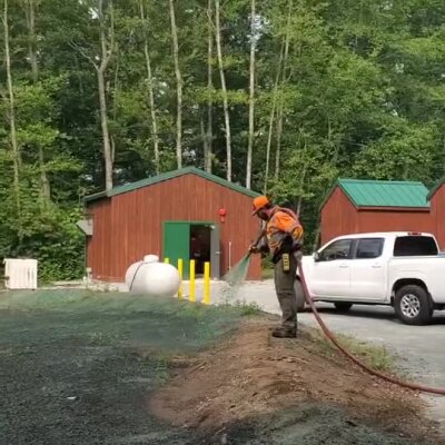 Worker spraying erosion control mulch near animal shelter buildings and parked white truck.
