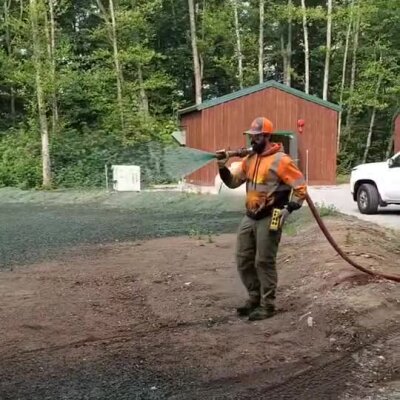 Worker spraying erosion control hydroseed on a bare soil area near wooded shelter.