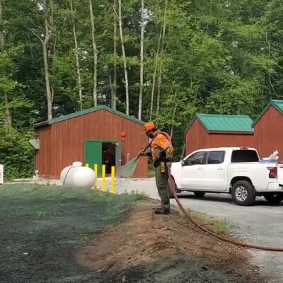 Worker spraying erosion control mulch near animal shelter buildings in wooded area.