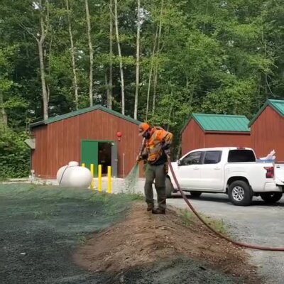 Worker spraying erosion control material near animal shelter buildings in wooded area.