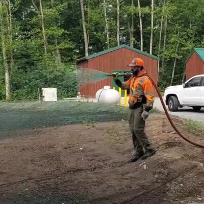 Worker spraying erosion control hydroseed on soil near buildings and forest.