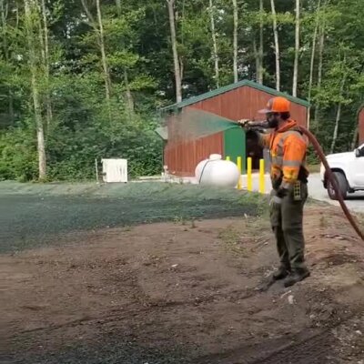 Worker spraying erosion control mulch on soil near wooded area and building.