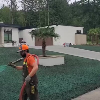 Technician spraying hydroseed on a new lawn in front of modern high-end home.