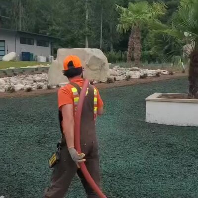 Worker applying hydroseed on residential front yard with palm trees and landscaping.