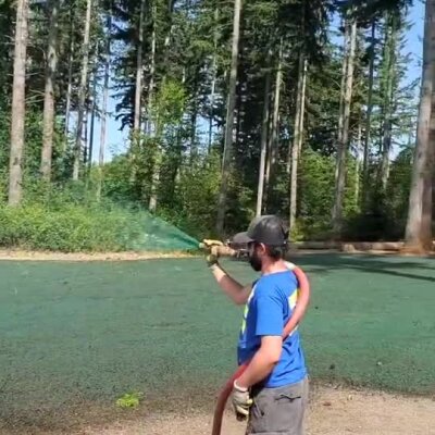 Technician spraying hydroseed over cleared soil near forested area under clear sky.