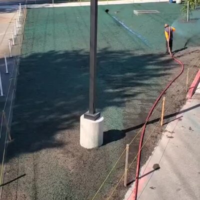 Hydroseeding worker spraying green seed mixture on school playground area with concrete and light pole.