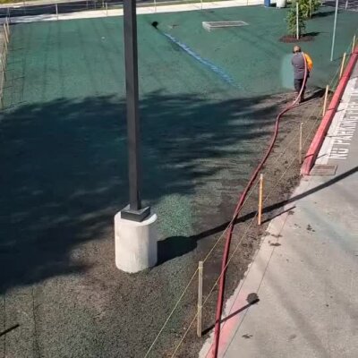 Worker applying hydroseeding on a large green field near a streetlight pole, sunny day Bothell.